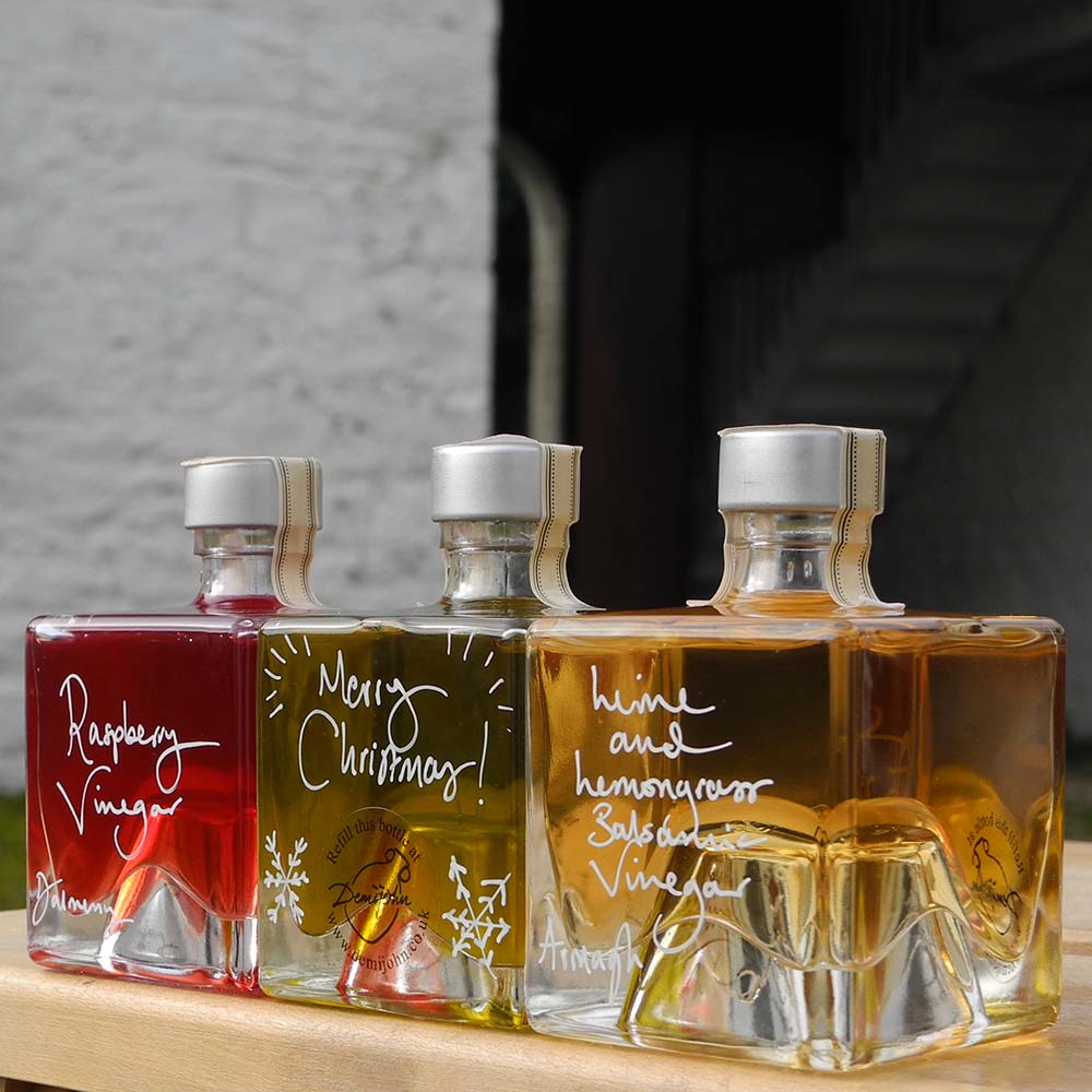 Three Mystic 100ml stacking bottles containing Lime and Lemongrass Balsamic Vinegar, Spinelli Tuscan Extra Virgin Olive Oil and Raspberry Vinegar. The labels are handwritten in white pen in front of the red Demijohn shutters at Glenlair Steading. there is a personalised message which says Merry Christmas