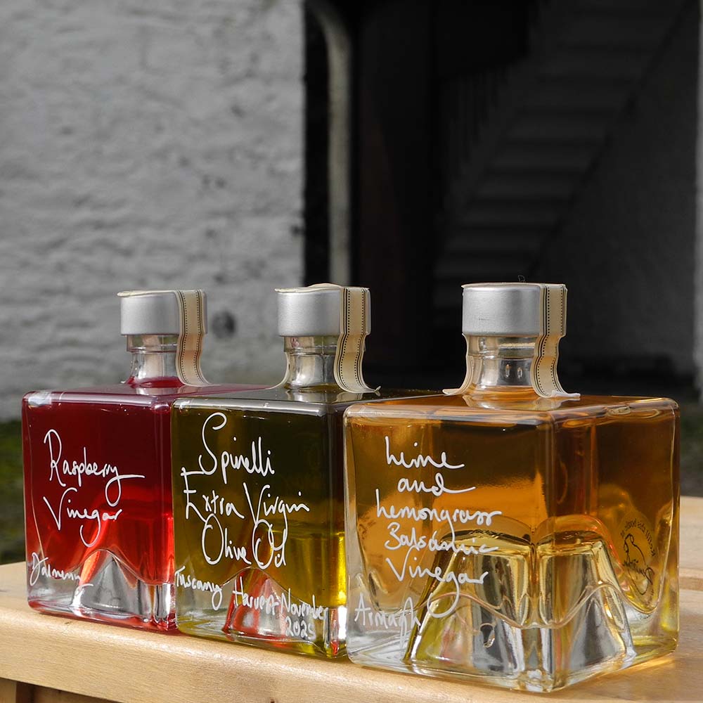 Three Mystic 100ml stacking bottles containing Lime and Lemongrass Balsamic Vinegar, Spinelli Tuscan Extra Virgin Olive Oil and Raspberry Vinegar. The labels are handwritten in white pen in front of the red Demijohn shutters at Glenlair Steading. 
