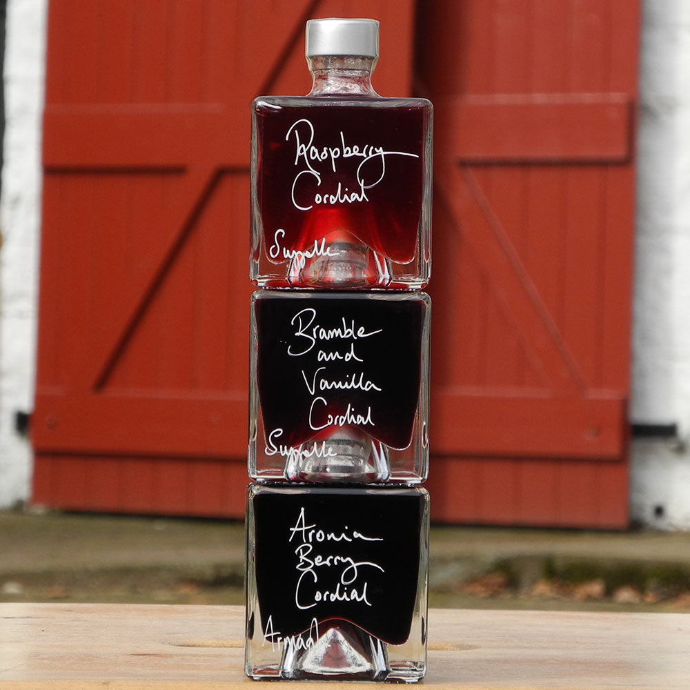 A gift set of three cube shaped, 100ml glass bottles stacked vertically on a wooden surface, each filled with a different handmade alcohol free drink: Raspberry Cordial, Bramble and Vanilla Cordial, and Aronia Berry Cordial, with white handwritten labels. A red barn door is in the background.