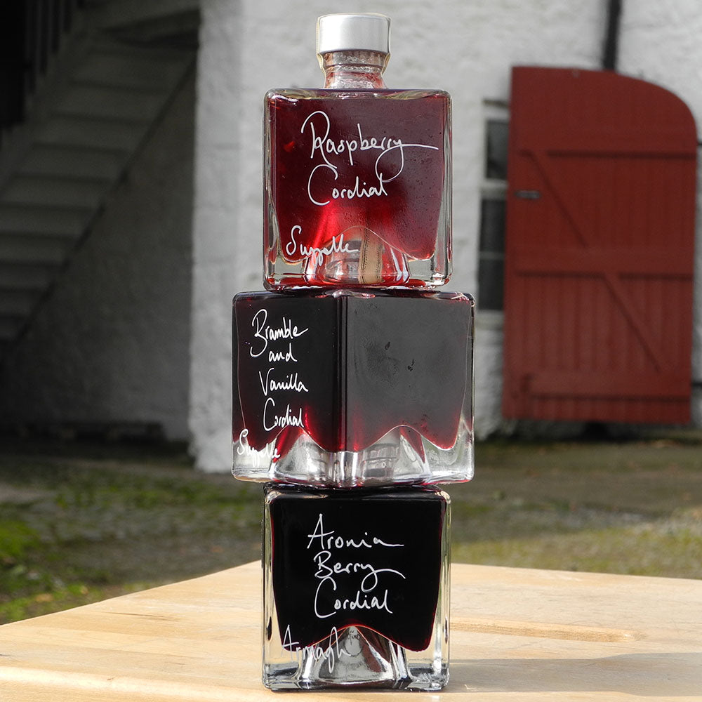 A gift set of three cube shaped, 100ml glass bottles stacked vertically on a wooden surface, each filled with a different handmade alcohol free drink: Raspberry Cordial, Bramble and Vanilla Cordial, and Aronia Berry Cordial, with white handwritten labels. A red barn door is in the background.