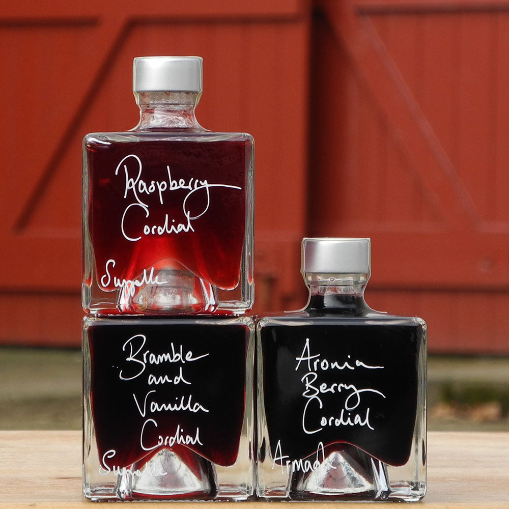 A gift set of three cube shaped, 100ml glass bottles stacked vertically on a wooden surface, each filled with a different handmade alcohol free drink: Raspberry Cordial, Bramble and Vanilla Cordial, and Aronia Berry Cordial, with white handwritten labels. A red barn door is in the background.