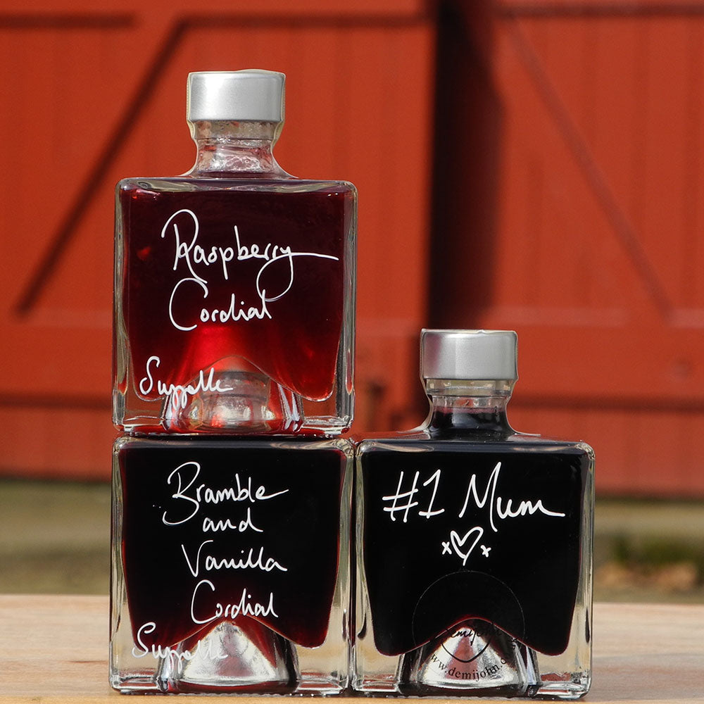 A gift set of three cube shaped, 100ml glass bottles stacked vertically on a wooden surface, each filled with a different handmade alcohol free drink: Raspberry Cordial, Bramble and Vanilla Cordial, and Aronia Berry Cordial, with white handwritten labels. One bottle has a handwritten message #1 Mum and a loveheart. A red barn door is in the background.