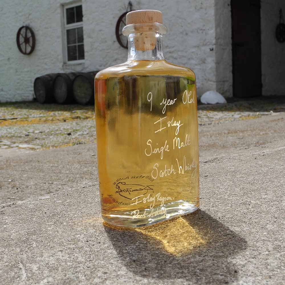 Hipflask shaped glass bottle of 9 year old Islay Single Malt Scotch Whisky on a concrete surface with traditional buildings in the background.