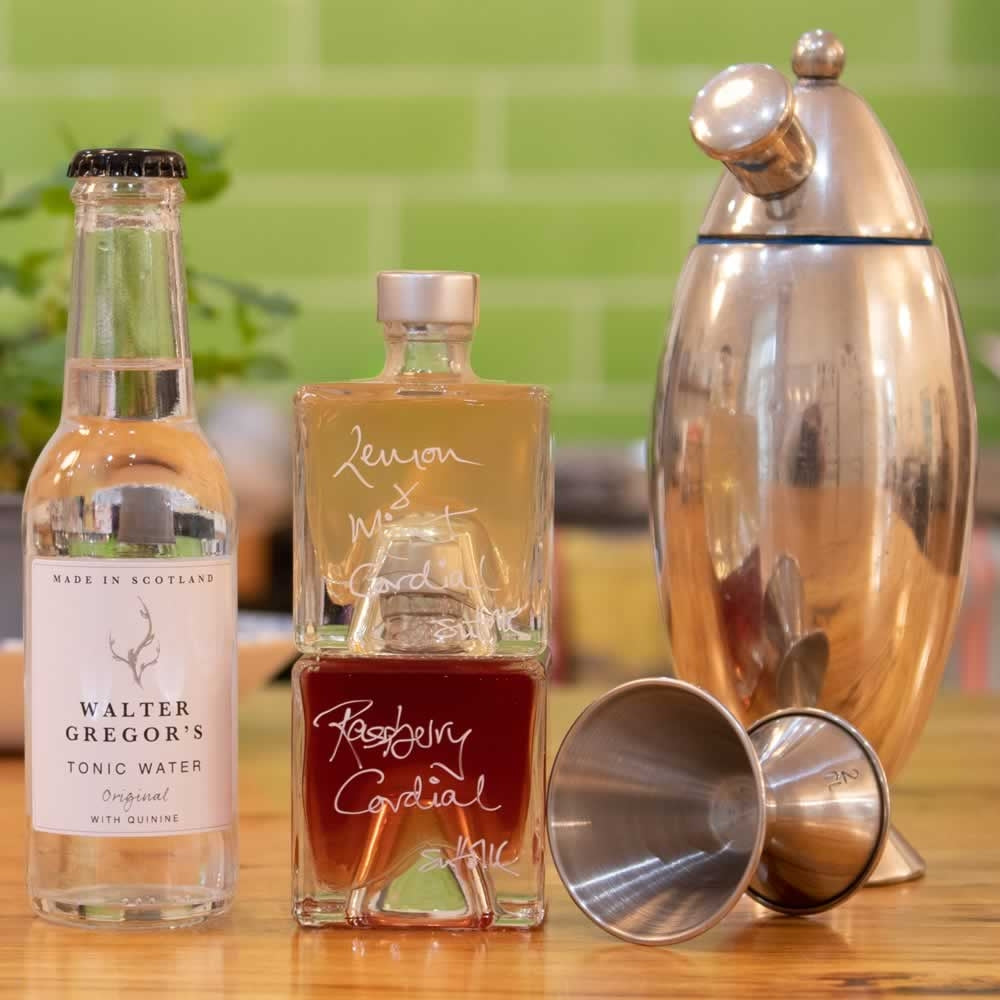 Mummy's Raspberry Fresh Mocktail Gift Set. Two cube shaped 100ml stacking bottles of different handmade cordials stacked on a table next to a bottle of tonic and a cocktail shaker. background.